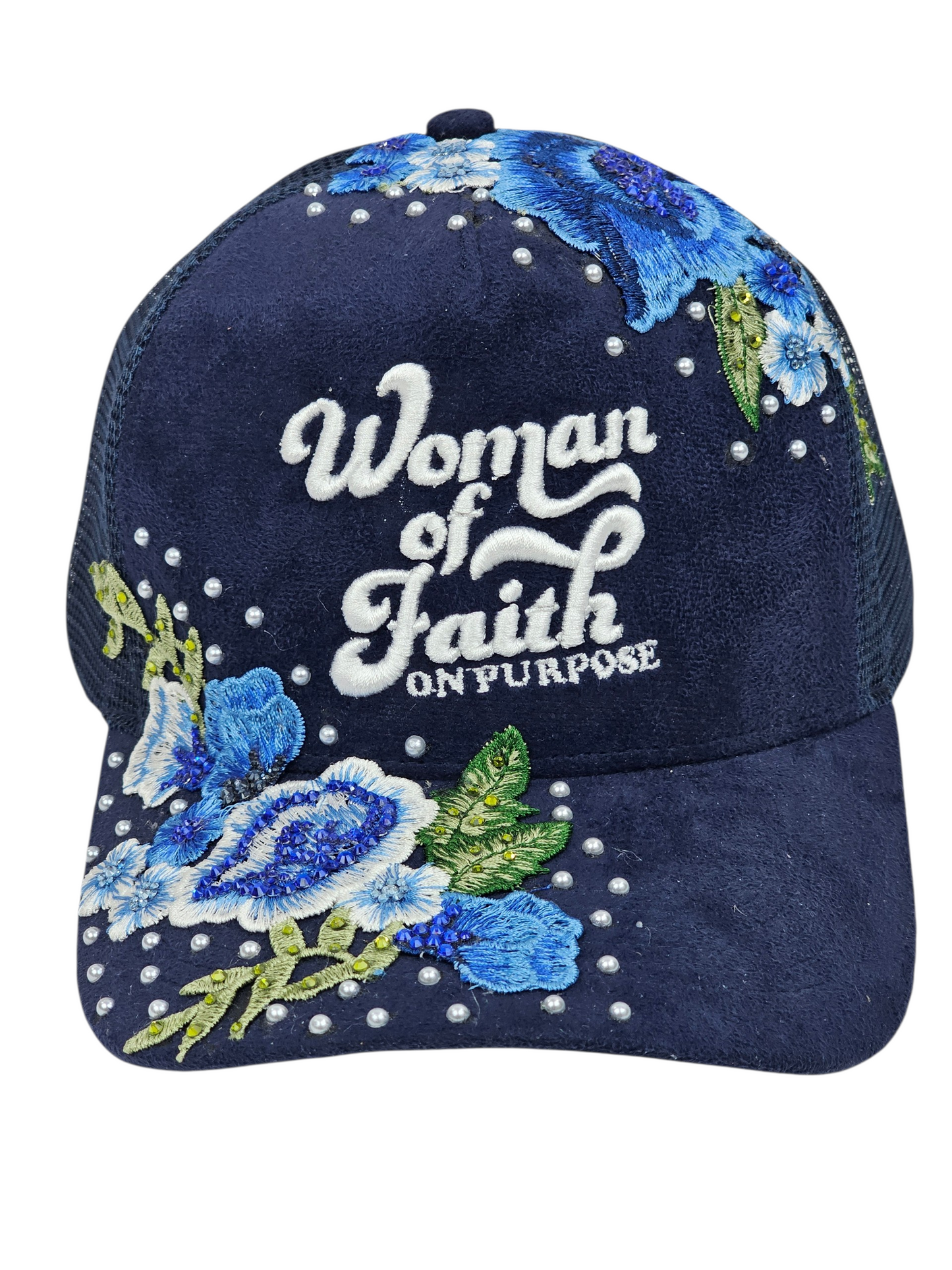 A navy blue trucker-style cap with embroidered blue and white flowers, green leaves, and white rhinestones. The front features the embroidered text 'Woman of Faith ON PURPOSE'.