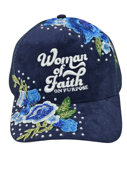 A navy blue trucker-style cap with embroidered blue and white flowers, green leaves, and white rhinestones. The front features the embroidered text 'Woman of Faith ON PURPOSE'.
